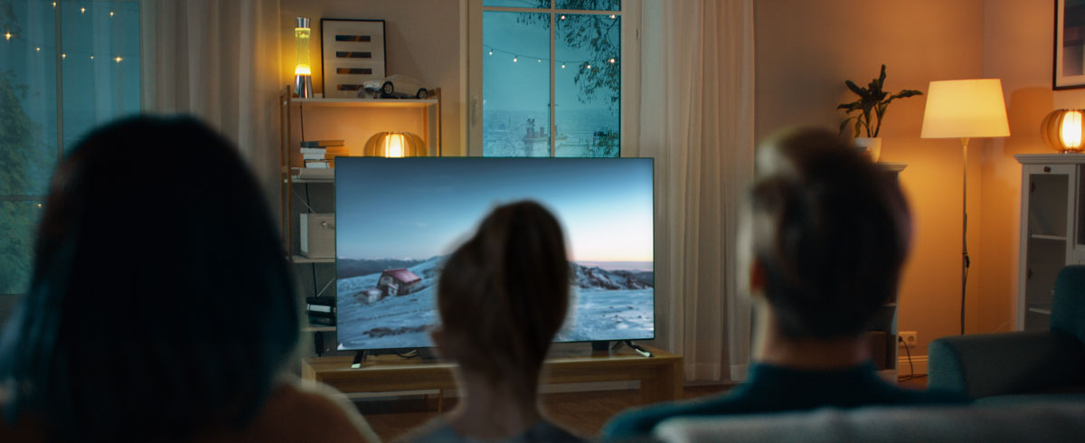 Eine Familie sitzt zusammen im Wohnzimmer und schaut gemeinsam auf einem gro&szlig;en Fernseher fern